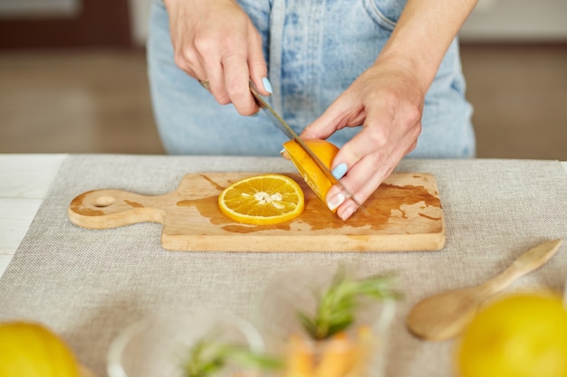 Фото Женская рука режет апельсин, женщина готовит, делает свежий лимонад из цитрусовых и розмарина в стакане на белом столе дома, летний напиток, очищает здоровую воду.