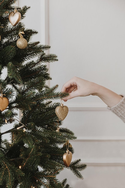 Female hand decorate beautiful Christmas tree with golden baubles and toys in front of white wall Elegant aesthetic Christmas New Year holidays celebration preparation background