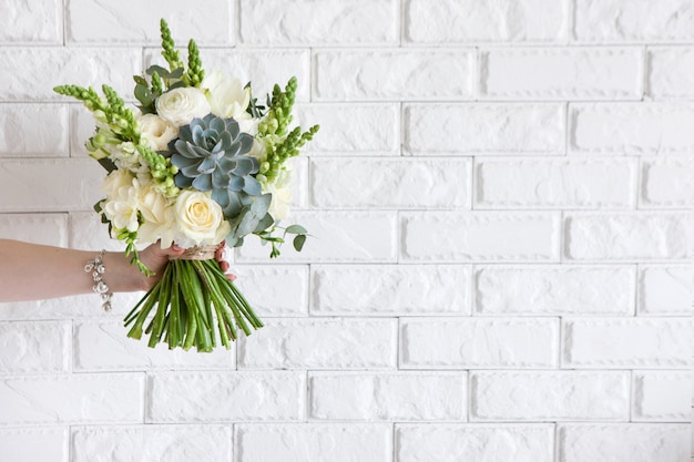 여성 손 장미와 흰색 벽돌 배경에 즙을 제공합니다. 어머니 또는 여성을위한 선물, 꽃집 작업, 웨딩 장식, 아름다운 꽃다발 판매 개념