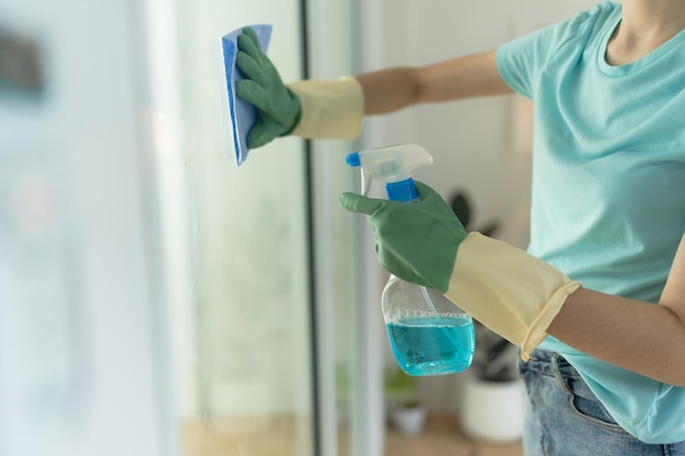 Female hand in green gloves cleaning window with blue rag and spray detergent