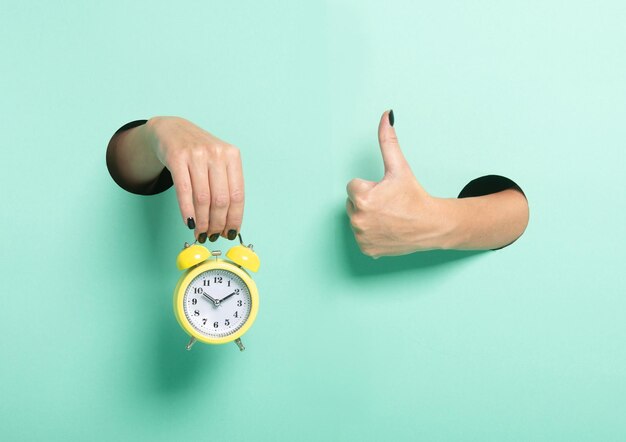 Female hand hold alarm clock and show tumb sign through a hole on neon mint background Just in time concept