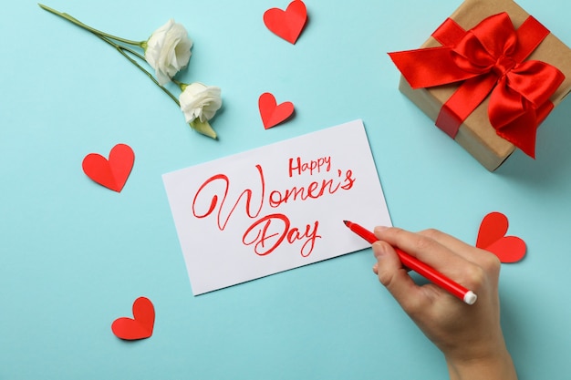 Female hand hold felt - tip pen, text Happy Women's Day, gift box, roses and hearts on blue background