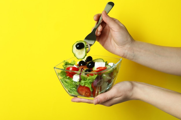 Female hand hold fork and bowl of greek salad on yellow background