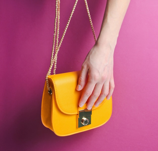 Женская рука держит и открывает модную желтую кожаную сумку с золотой цепочкой на фиолетовом фоне.