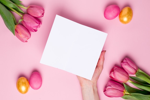 Female hand hold present card on a pink surface with flowers and easter eggs