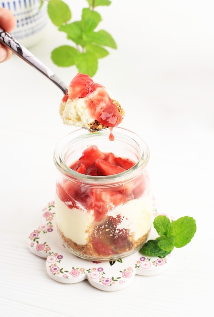 Female Hand Hold Spoon with Traditional Italian Dessert Vanilla Strawberry Panna Cotta in a Vintage Jars with Fresh Berries on a White Wooden Background.