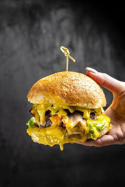 Фото Женская рука держит большой аппетитный укушенный бургер на темном бетонном фоне концепция фаст-фуда и нездоровой пищи вертикальное место изображения для текста