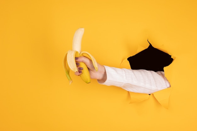 Женская рука, держащая банановый фрукт, концепция питания, человеческая рука, держащая банан в отверстии оранжевого фона
