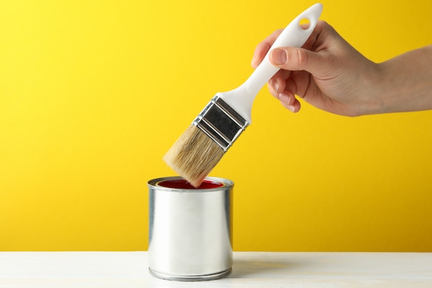 Female hand holding brush over the can with red paint against yellow background