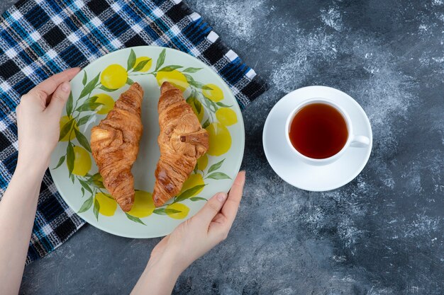 お茶のカップの近くに焼きたてのクロワッサンのカラフルなプレートを持っている女性の手