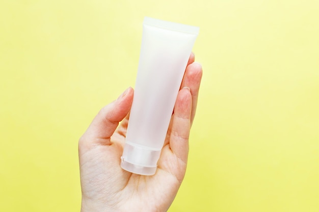 Female hand holding cream tube cosmetic products on a yellow background.