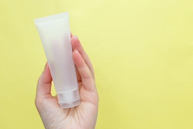 Female hand holding cream tube cosmetic products on a yellow background.