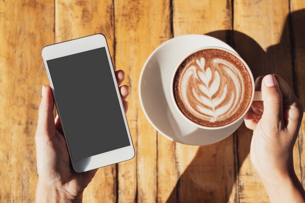 Female hand holding cup of hot cocoa or chocolate while holding mobile phone on wooden table, close up