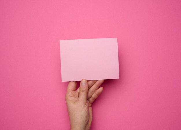 Female hand holding empty pink paper on a pink background. Copy paste image or text, close up