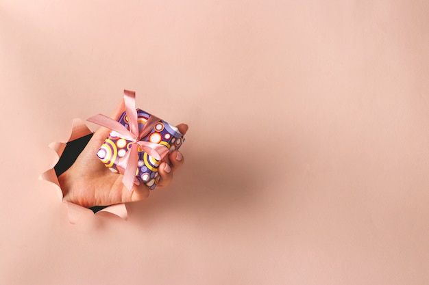 Женская рука держа подарочную коробку через круглое отверстие в розовой бумажной предпосылке, дне подарков, планировании подарка, космосе экземпляра
