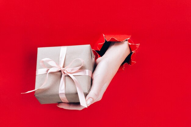 Female hand holding gift box wrapped in craft paper with pink ribbon through a hole in red.