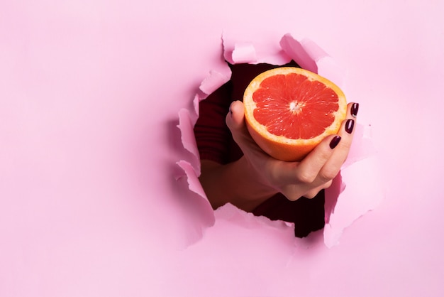 Female hand holding half of grapefruit through torn pink paper background.