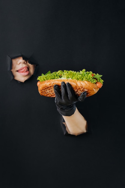 Female hand holding hot dog and looks through holes in black paper background street food concept