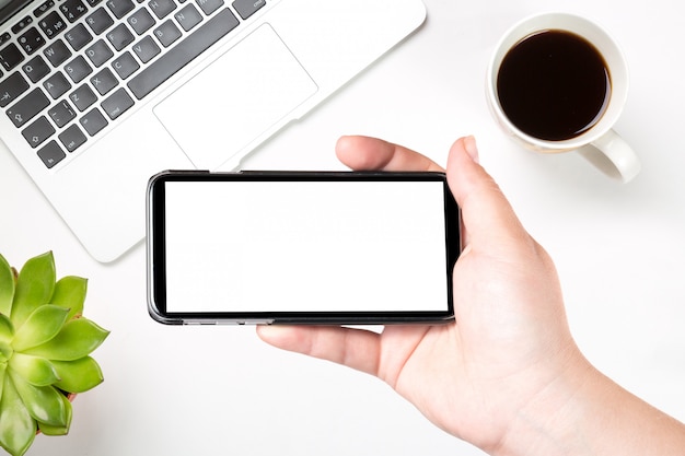 Female hand holding mobile smart phone with blank white screen against modern laptop, coffee and green plant.