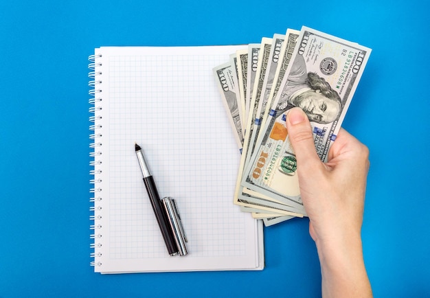 Female hand holding money above empty notepad with pen Business background Top view