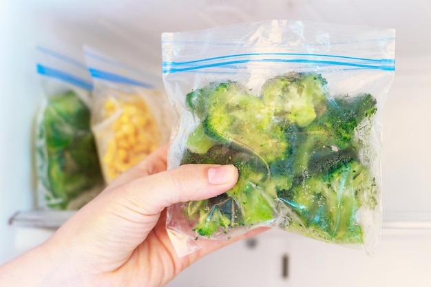 Foto mano femminile che tiene un pacchetto di broccoli congelati sullo sfondo del congelatore.