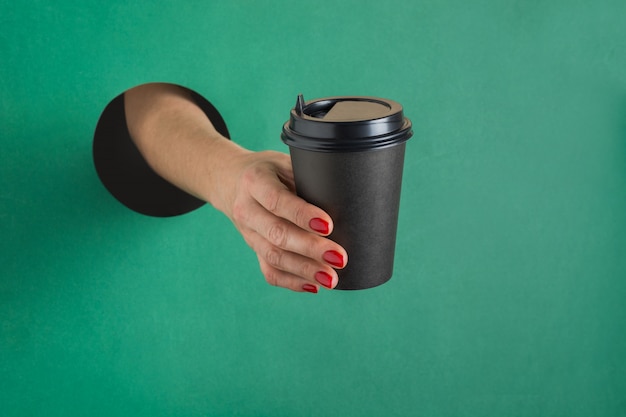 Female hand holding paper coffee cup isolated round hole in green paper.