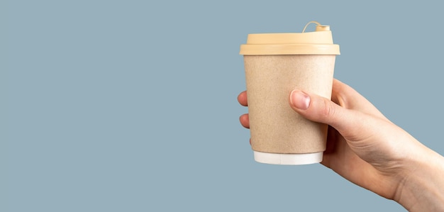 Female hand holding a paper cup of coffee over pastel blue background banner copy space Disposable takeaway takeout mug of latte with lid cover