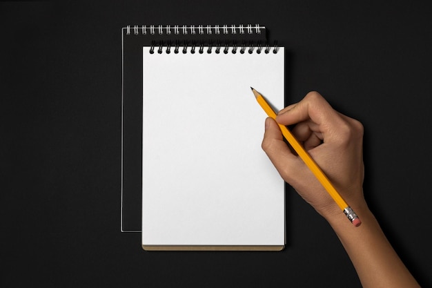 Female hand holding pencil over blank notebook on black top view