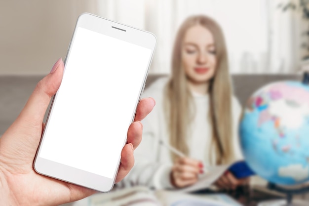 Female hand holding a phone with a blank screen on a background with a student girl teaches lessons at the table