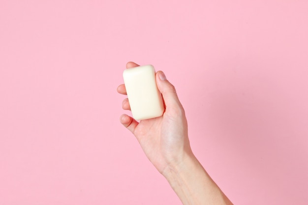 Female hand holding piece of soap against pink.