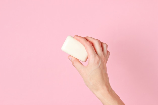 Female hand holding piece of soap against pink.