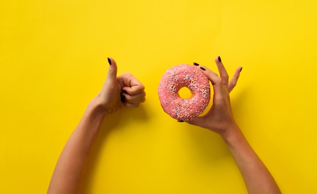 Female hand holding pink donut and showing like, ok sign over yellow background.