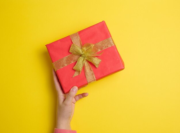 Female hand holding red gift box on a yellow background concept of congratulations on birthday