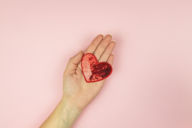 Female hand holding red sequin heart on pink background.