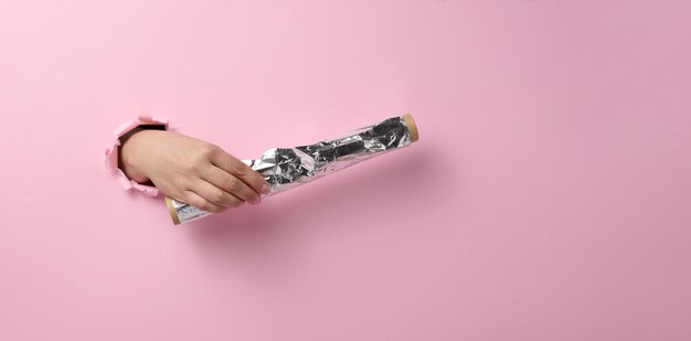 Female hand holding a scroll of gray food foil on a pink background, part of the body sticking out of a torn hole. Copy space, banner