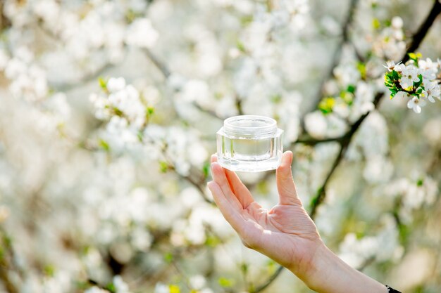 Female hand holding skin care cream near flowering tree. Spring season