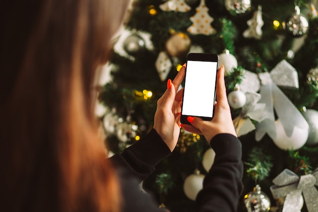 Female hand holding smartphone with blank white screen on of christmas tree