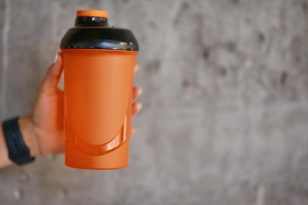 Female hand holding sports bottle with water against grey wall at gym selective focus