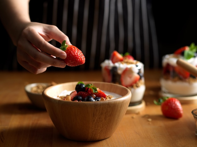 female hand holding strawberry adding on a bowl of granola with Greek yogurt and berries