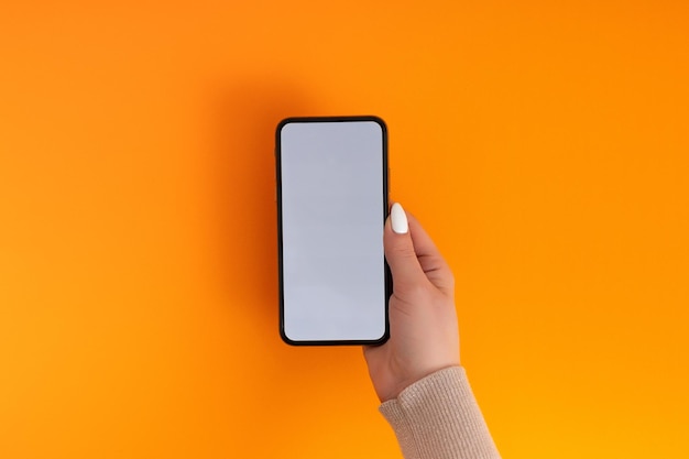 Женская рука держит и прикасается к мобильному смартфону с белым экраном Изолированный на оранжевом Фотошаблон для любых изображений на дисплее мобильного телефона Макет с легко снимаемым фоном монитора телефона