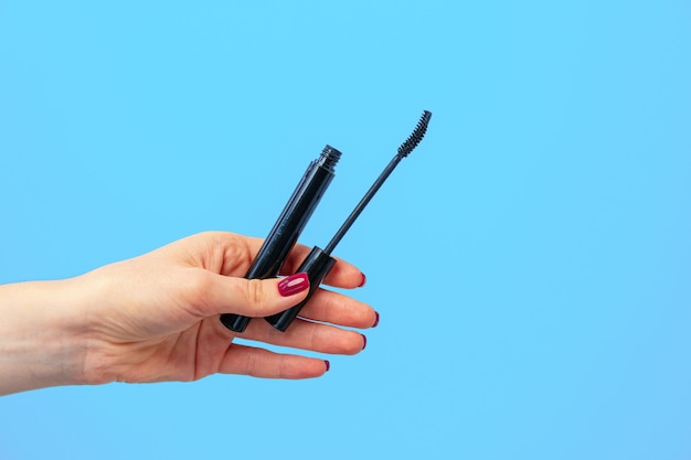 Female hand holding tube of mascara against blue close up