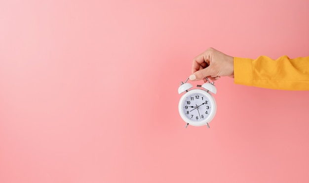 Female hand holding white alarm clock on pink background