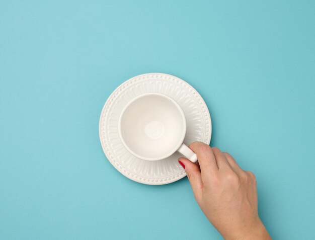 Photo female hand holding a white ceramic mug with a saucer on a blue background top view person