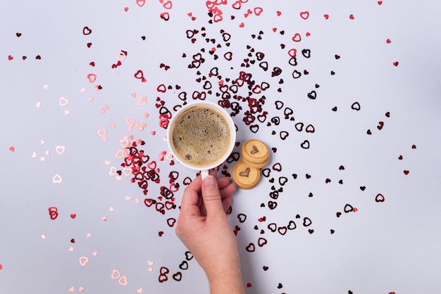 Female Hand Holding White Cup of Coffee Two Cookies Blue Background with Red Confetti