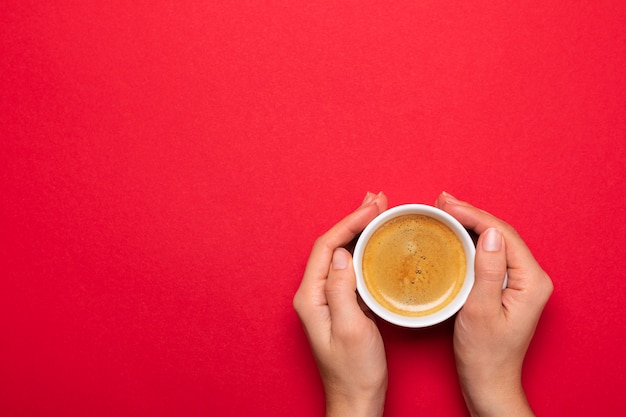 Female hand holding a white cup with black coffee on a red surface.