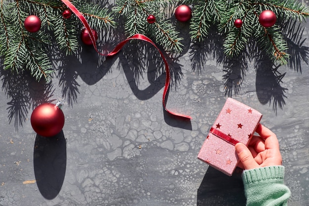 Женская рука держа завернутый Рождественский подарок с красной безделушка. Рождественская квартира лежала на абстрактной жидкой акриловой живописи. Пограничная гирлянда из натуральных еловых веточек и красных шаров с длинными тенями.