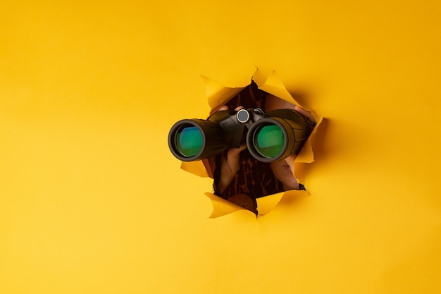 Female hand holds black binoculars on a yellow background. Journey, find and search concept. Banner.