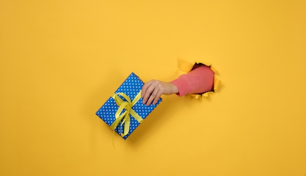 Female hand holds a box with a gift on a yellow background part of the body sticks out of