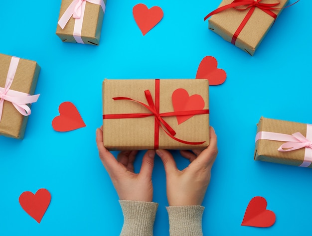 Female hand holds a box wrapped in brown kraft paper and tied with a silk ribbon on a blue background