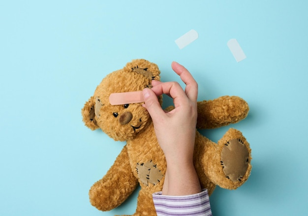 Female hand holds a brown teddy bear and glues a medical adhesive plaster on a blue background, tram treatment